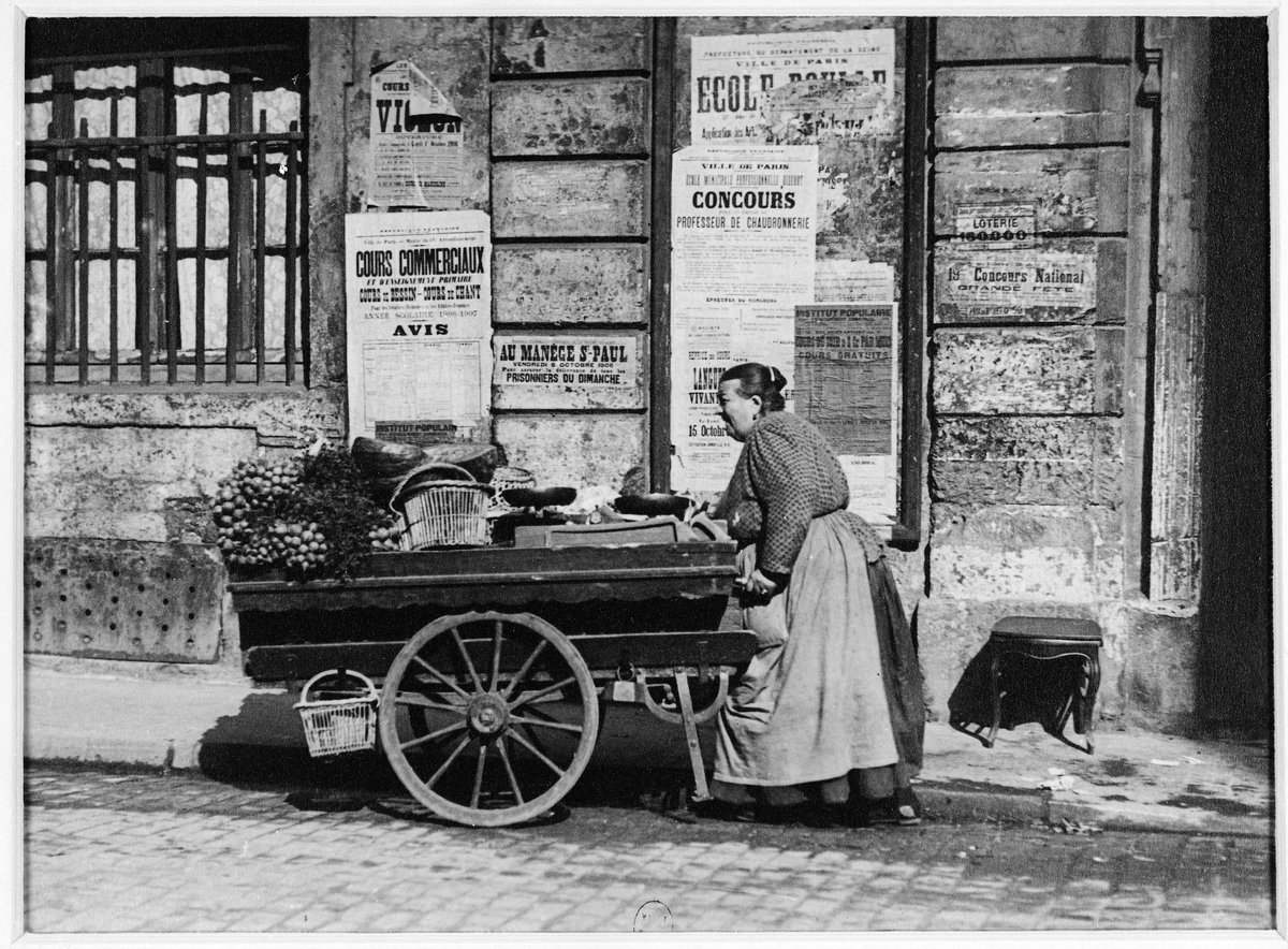 Photographie de 1906 : une marchande des quatre saisons pousse sa charrette de légumes dans une rue de Paris